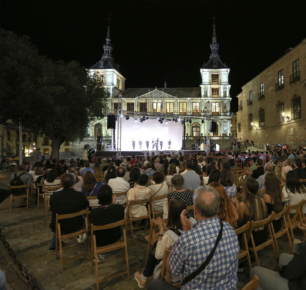 Un grupo de personas sentadas en sillas de madera observa un espectáculo de baile de noche en la plaza del Ayuntamiento de Toledo.